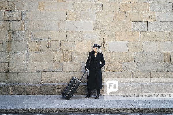 Reife Geschäftsfrau mit schwarzem Mantel und Ledermütze steht mit rollendem Koffer auf dem Bürgersteig