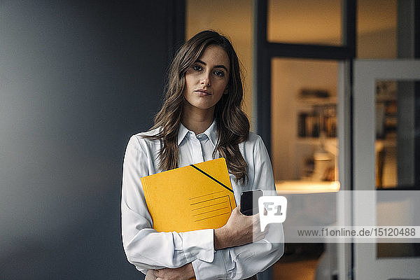 Portrait of serious young businesswoman holding folder and cell phone