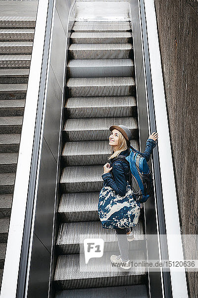 Smiling woman with backpack and travelling bag standing on escalator looking around
