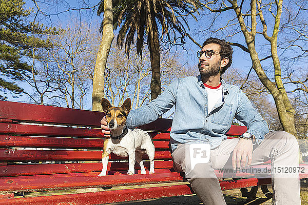 Zufriedener junger Mann sitzt auf Parkbank und streichelt seinen Hund