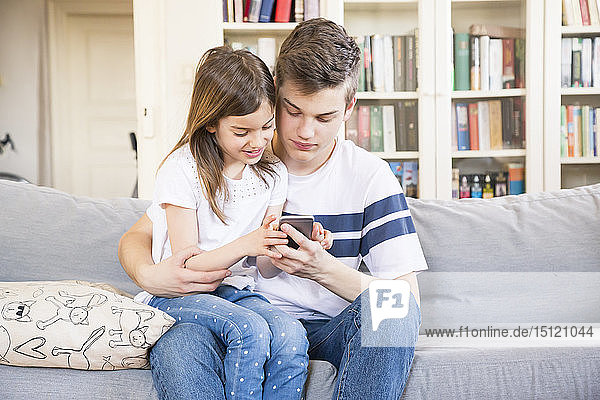 Teenager  der mit seiner kleinen Schwester zu Hause auf der Couch sitzt und auf sein Handy schaut