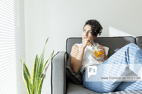 Entspannte junge Frau sitzt mit einem Erfrischungsgetränk auf dem Sofa