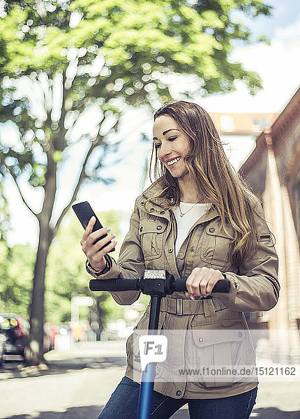 Porträt einer lächelnden Frau mit E-Scooter  die auf ein Smartphone schaut