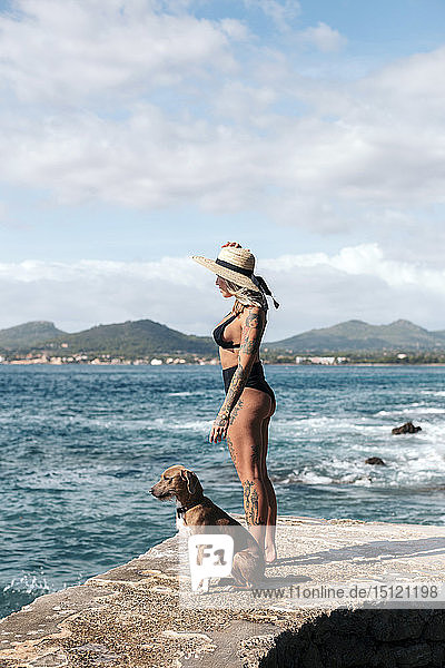 Spanien  Mallorca  tätowierte Frau steht neben ihrem Hund und schaut auf das Meer