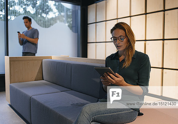 Junge Frau sitzt auf Couch mit Tablette mit Mann im Hintergrund