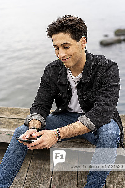Dänemark  Kopenhagen  lächelnder junger Mann sitzt am Wasser und benutzt sein Handy