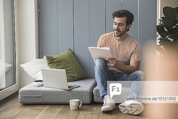 Junger Mann sitzt auf Matratze  benutzt Laptop und macht Notizen