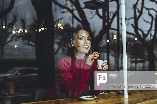 Smiling young woman with cup of coffee behind windowpane in a cafe