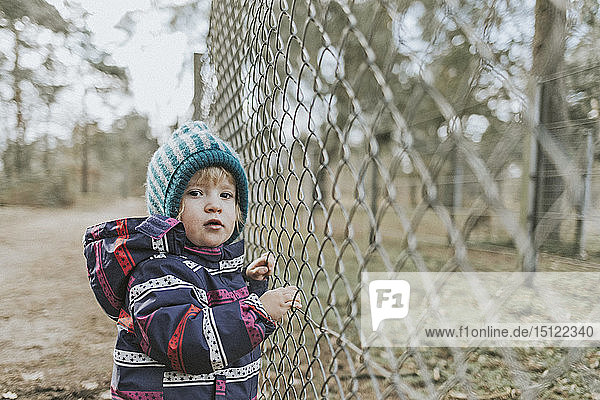 Portrait of toddler girl in warm clothes at a fence in forest
