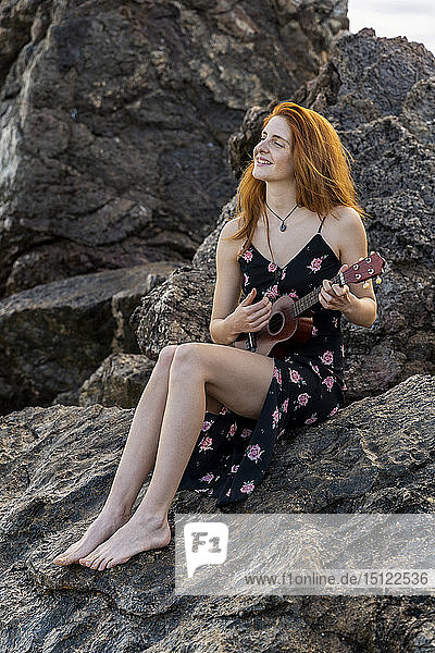 Lächelnde rothaarige junge Frau sitzt auf einem Felsen und spielt Ukulele