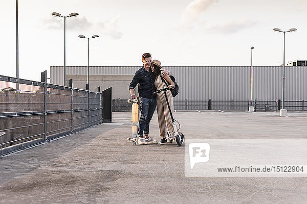 Junges Paar mit Longboard und Elektroroller umarmt sich auf dem Parkdeck