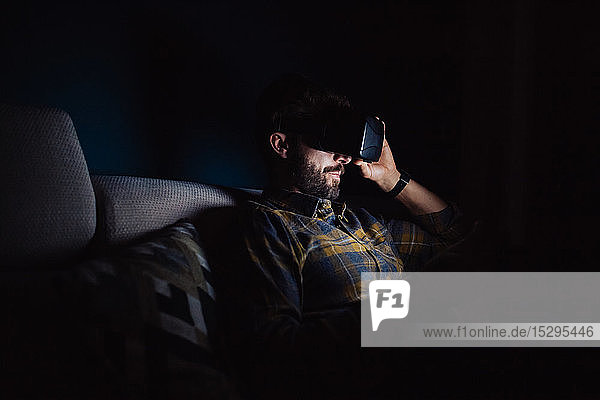 Bärtiger mittelgroßer Mann  der im Dunkeln auf dem Sofa sitzt und durch ein Virtual-Reality-Headset nach unten blickt