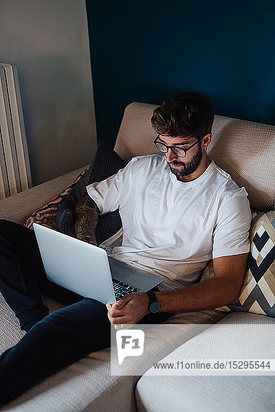 Mittelgroßer erwachsener Mann  der sich mit einem Laptop auf dem Sofa zurücklehnt