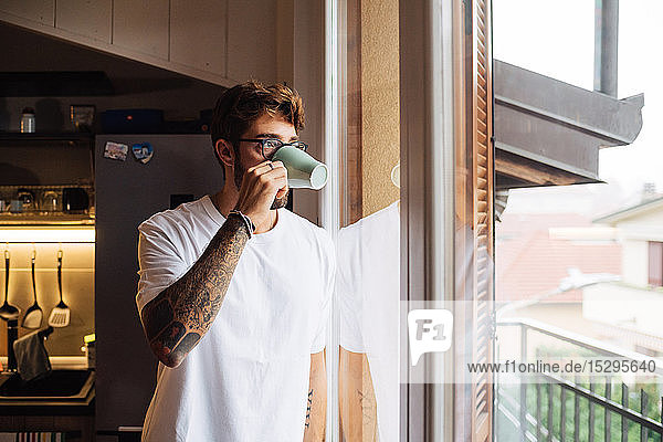 Mittelgroßer erwachsener Mann trinkt Kaffee  während er durch ein Wohnungsfenster hinausschaut
