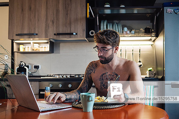 Mittelgroßer erwachsener Mann mit Tätowierungen  der Omelett isst  während er am Laptop tippt
