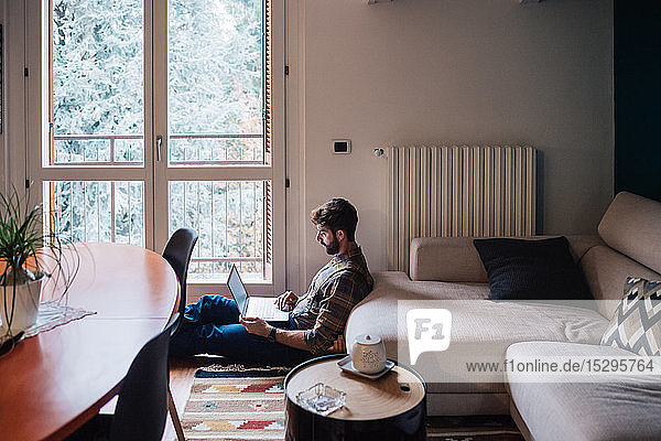 Mittelgroßer erwachsener Mann sitzt mit Laptop auf dem Wohnzimmerboden