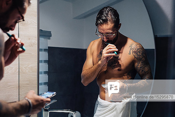 Mittelgroßer erwachsener Mann mit Tätowierungen  der sich die Zähne putzt und sein Smartphone am Badezimmerspiegel betrachtet  Spiegelbild