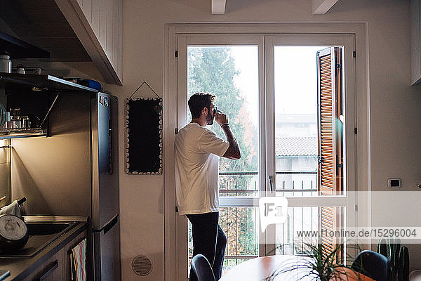 Mittelgroßer erwachsener Mann hält Kaffee trinkend in der Hand und schaut durch das Wohnungsfenster hinaus