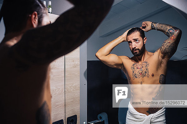 Mittelgroßer erwachsener Mann mit Tätowierungen  der am Badezimmerspiegel die Haare kämmt  Spiegelbild