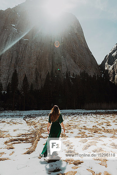 Young woman in long dress standing in sunlit snow covered mountain landscape rear view Yosemite Village California USA