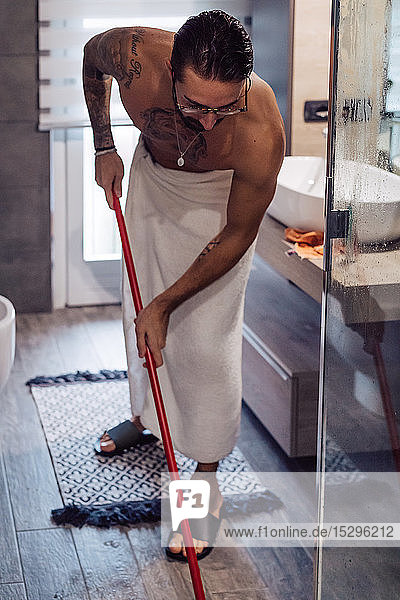 Mittelgroßer erwachsener Mann in Handtuch gewickelt reinigt Badezimmerboden