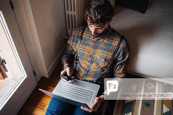 Mittelgroßer erwachsener Mann sitzt auf dem Wohnzimmerboden mit Laptop und Smartphone