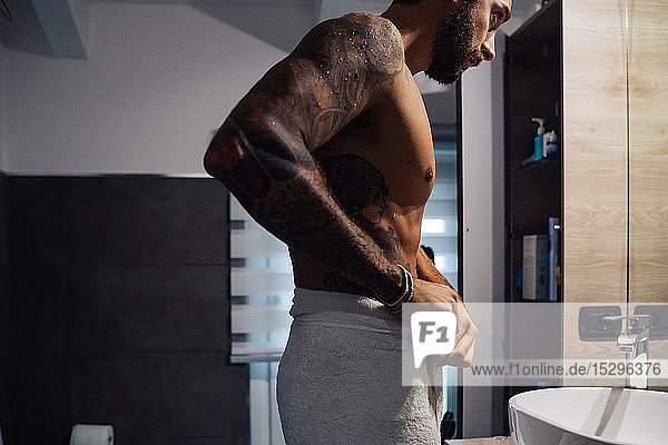 Mittelgroßer Mann mit Tätowierungen  der am Badezimmerspiegel ein Handtuch um die Taille legt