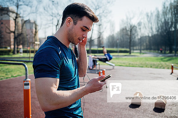 Gymnastik im Fitnessstudio im Freien  junger Mann hört Kopfhörer und schaut auf ein Smartphone