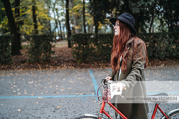 Young woman with long red hair pushing bicycle in autumn park