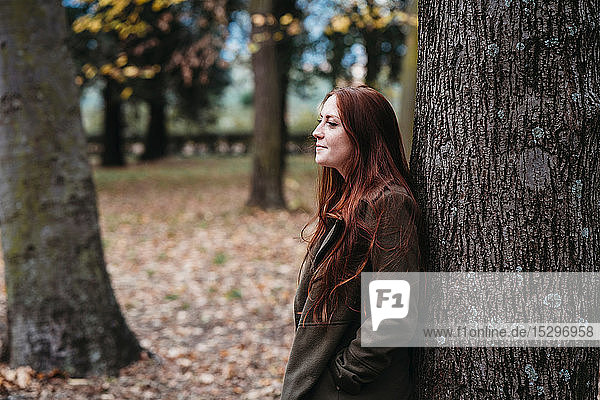 Junge Frau mit langen roten Haaren lehnt an Baumstamm im Herbstpark