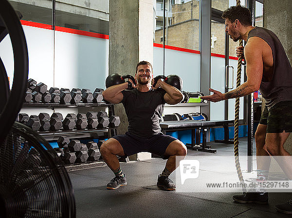 Man motivating friend doing squats  lifting kettlebells in gym