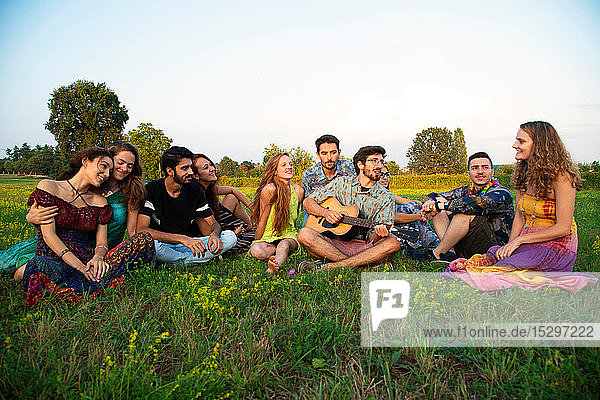 Large group of young adults sitting in field listening to acoustic guitar