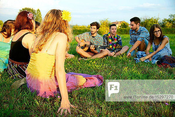 Große Gruppe junger Erwachsener sitzt im Feld und hört akustische Gitarre