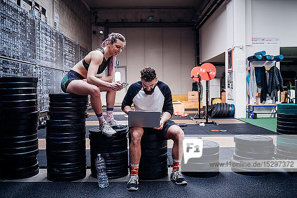 Junge Frau und Mann trainieren gemeinsam im Fitnessstudio  sitzen auf Gewichten und schauen auf Smartphone und Laptop