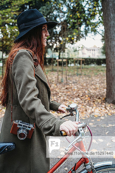 Young woman with long red hair pushing bicycle in autumn park
