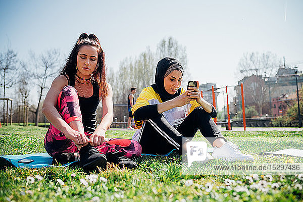 Calisthenics-Kurs im Fitnessstudio im Freien  junge Frauen sitzen auf Gras  binden Schnürsenkel und schauen auf das Smartphone