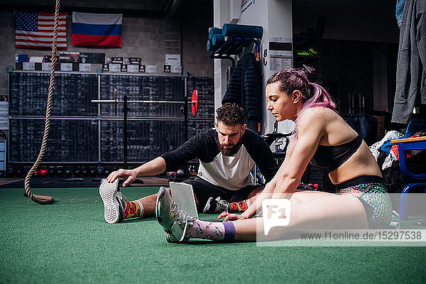 Junge Frau und Mann trainieren gemeinsam im Fitnessstudio  sitzen auf dem Boden  machen Aufwärmübungen und schauen auf den Laptop
