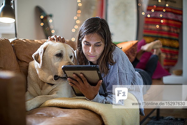 Labrador Retriever und junge Frau liegen auf einem Wohnzimmersofa und schauen auf ein digitales Tablett