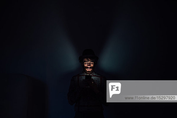 Bärtiger junger Mann benutzt Smartphone in dunklem Raum