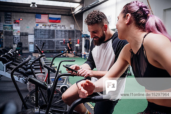 Junge Frau und Mann trainieren gemeinsam auf Fitness-Fahrrädern  Blick auf Smartphone