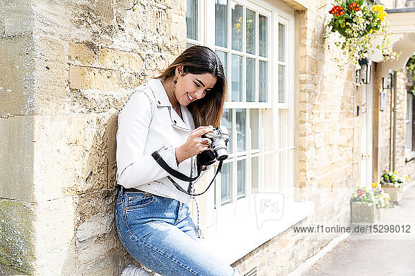 Junge Frau lehnt an Straßenmauer und betrachtet Fotos mit Digitalkamera  Cotswolds  England