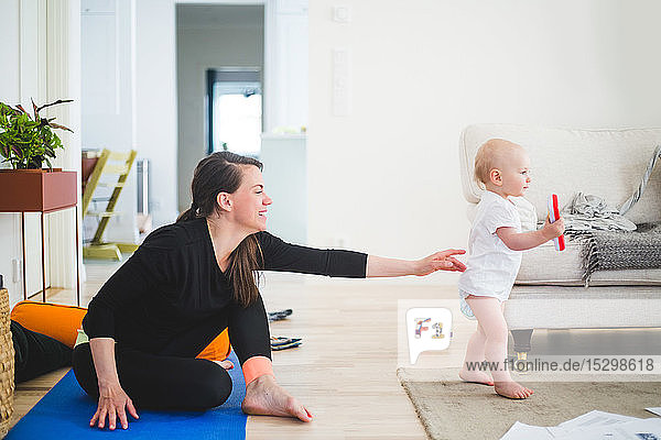Seitenansicht einer berufstätigen Mutter  die nach ihrer Tochter greift  während sie zu Hause im Wohnzimmer auf einer Gymnastikmatte sitzt