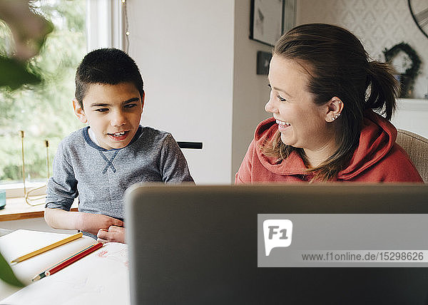 Lächelnde Mutter sieht autistischen Sohn an  der sich im Wohnzimmer ein Video über einen Laptop ansieht