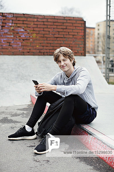 Ganzfigur eines lächelnden Teenagers mit einem Smartphone in der Hand  der in der Stadt auf der Straße sitzt