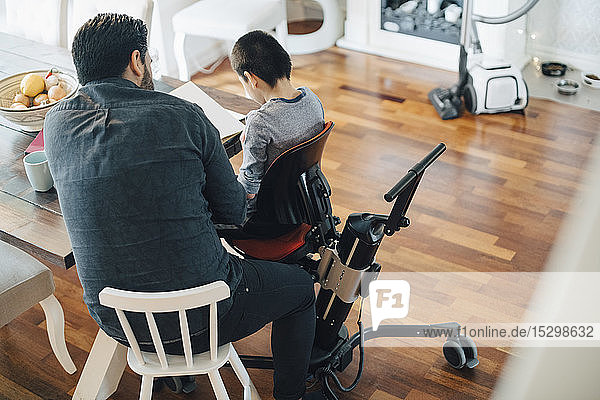 Hochwinkelaufnahme eines Vaters  der einen autistischen Sohn unterrichtet  der im Wohnzimmer auf einem Rollstuhl sitzt