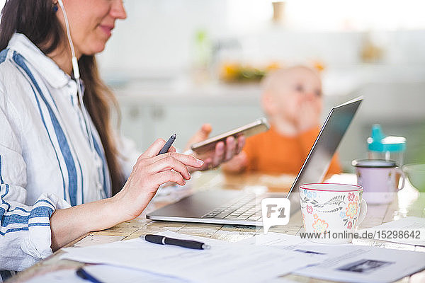 Midsection of working mother using technologies while daughter sitting in background at dining table