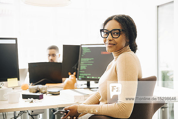 Portrait of female programmer sitting by desk with colleague in background at creative office