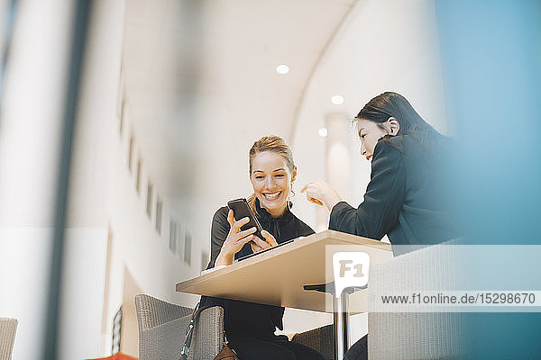 Niedriger Blickwinkel auf eine lächelnde Geschäftsfrau  die ihr Smartphone mit einer Tischkollegin im Büro teilt