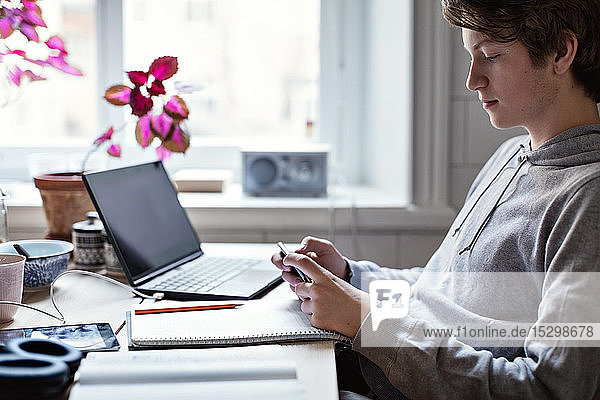 Seitenansicht eines Teenagers  der ein Mobiltelefon benutzt  während er zu Hause mit Büchern und Laptop auf dem Tisch sitzt