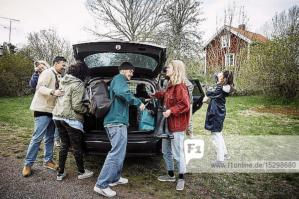 Smiling female and male friends removing luggage from car trunk on road against sky